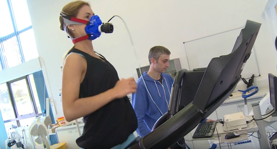 A researcher watches someone run on a treadmill, wearing an oxygen monitoring mask