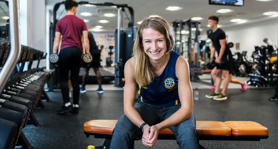 A student at the gym sits on a bench and smiles at the camera