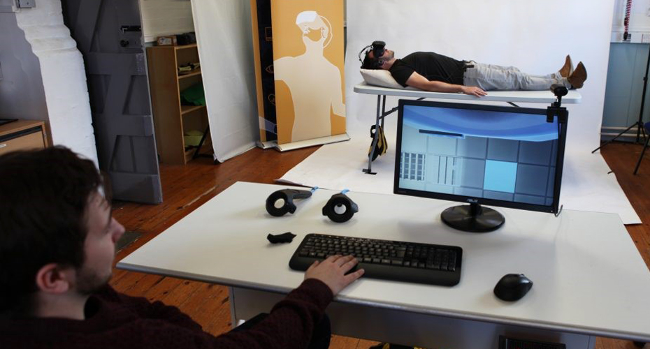 A participant lying on a bed wearing a VR headset, while someone else watches their view on a monitor