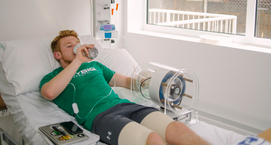 A student lying on a hospital bed, drinking water while his arm is immobilised as part of an experiment