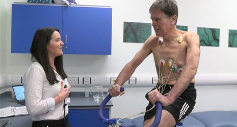 A researcher talking to an older man as part of an experiment. He is cycling, with electrodes on his chest linked to a machine.