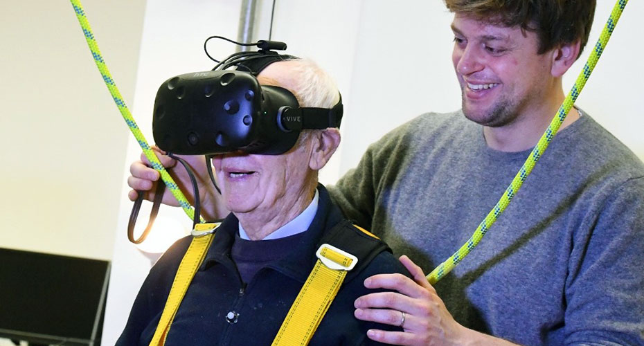An older man wearing a VR headset. A younger man supports his shoulders.