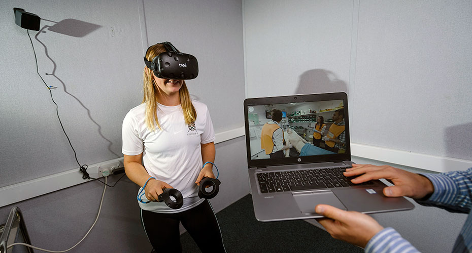 A student wearing a VR headset and holding controllers. On a laptop in front of her we see her moving in a VR environment.